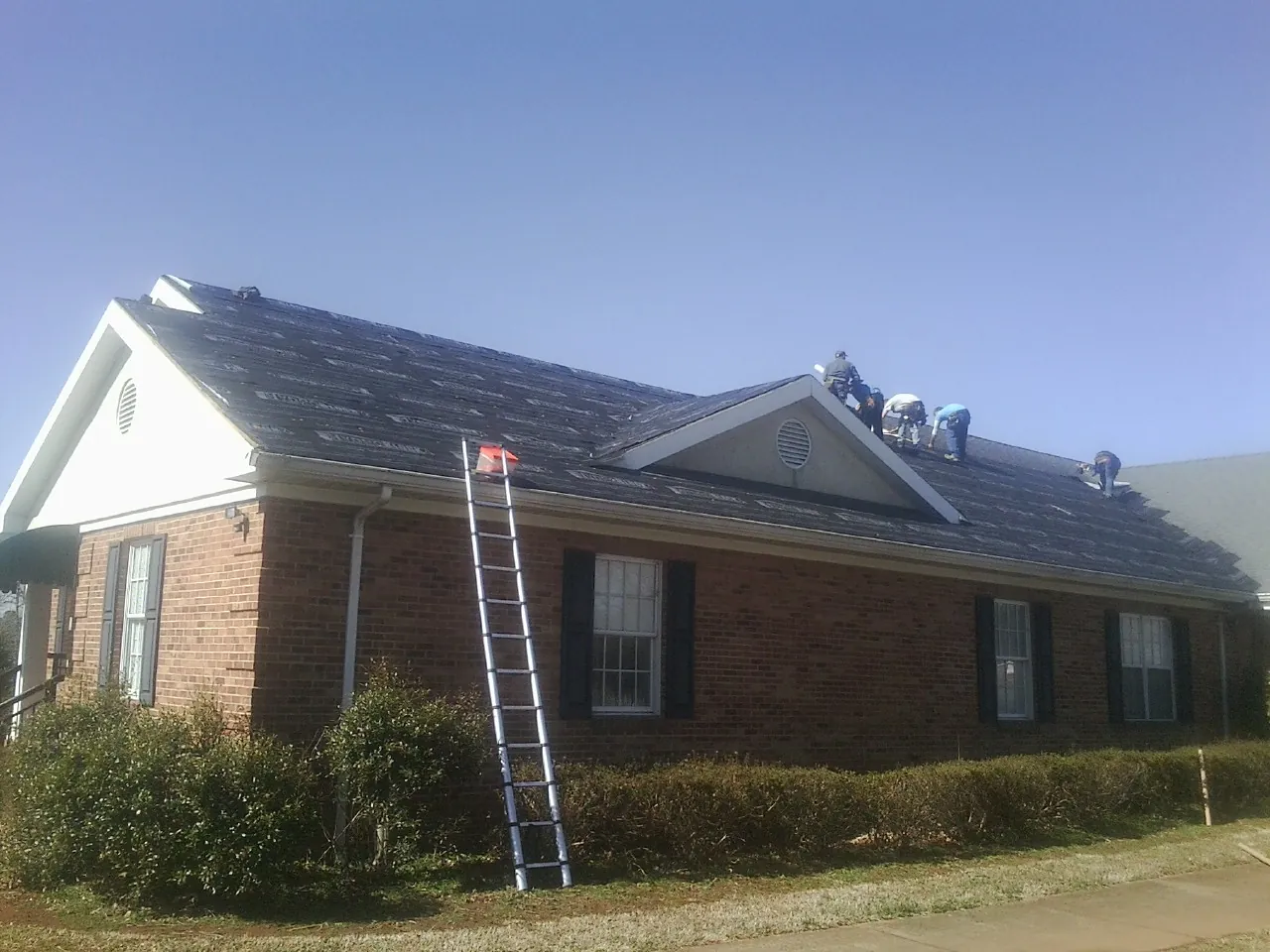 Crew tearing shingles off a brick ranch roof with a ladder set — Emergency Roof Repair work by our crew in Lempster