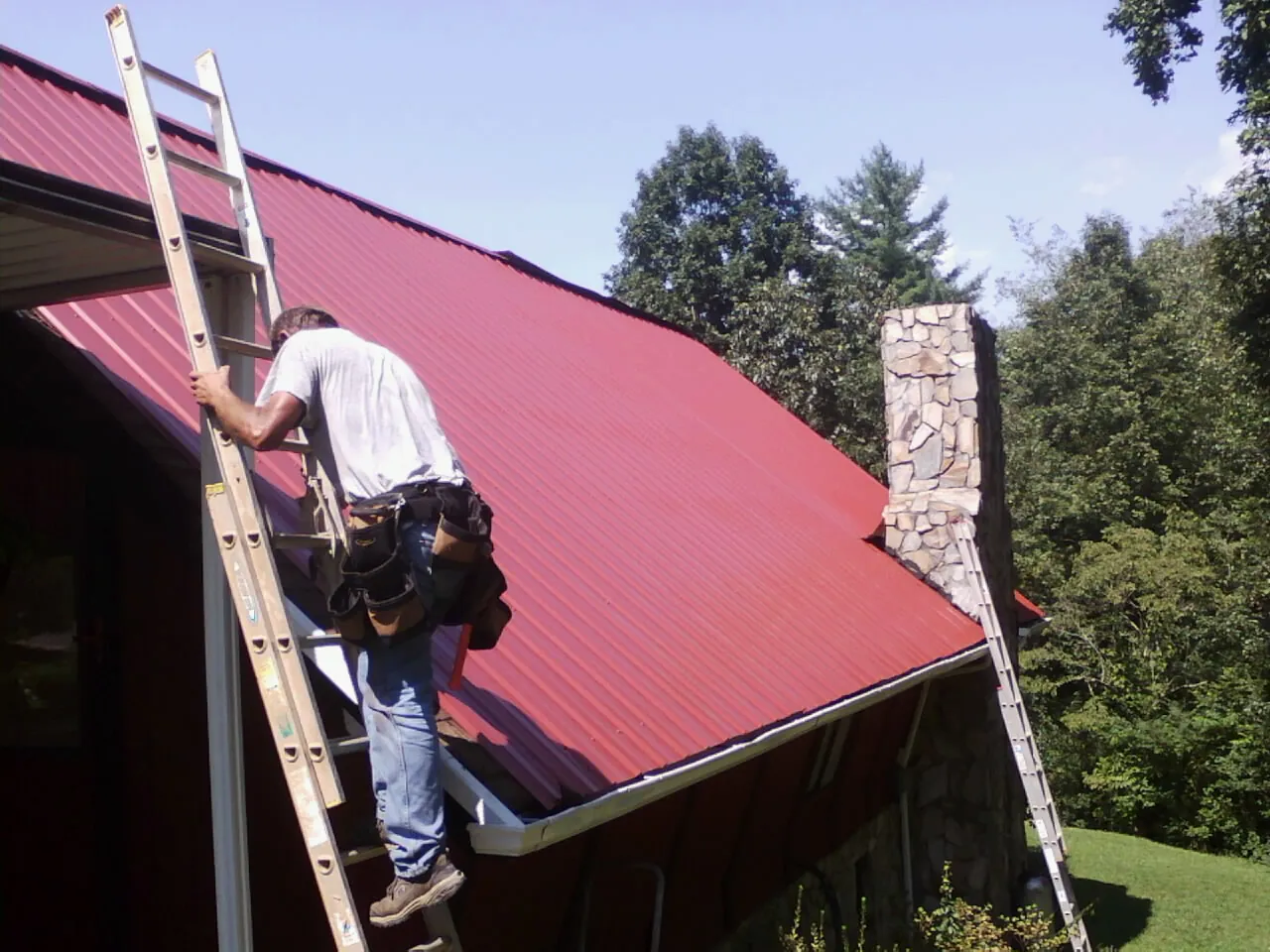 Roofer on a ladder beside a red metal roof and stone chimney — Soffit & Fascia Repair by Roofing Brothers in Lempster